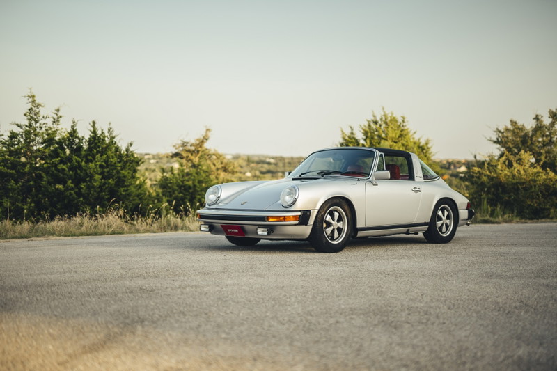 Silver 1977 Porsche 911S Targa with black roof, red interior, classic Fuchs rims, and black trim on an open road.
