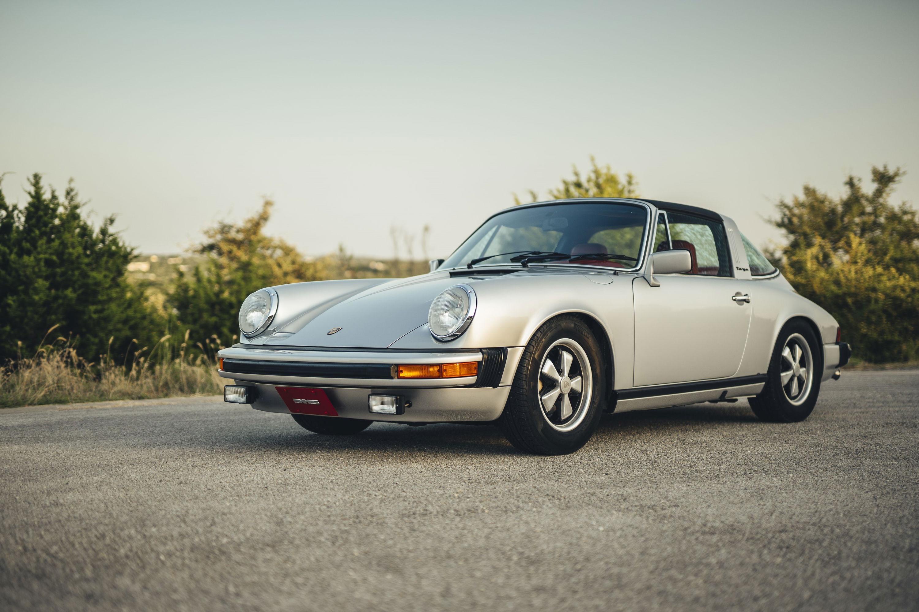 Silver metallic 1977 Porsche 911S Targa with black removable roof, iconic Fuchs rims, and vibrant red interior.
