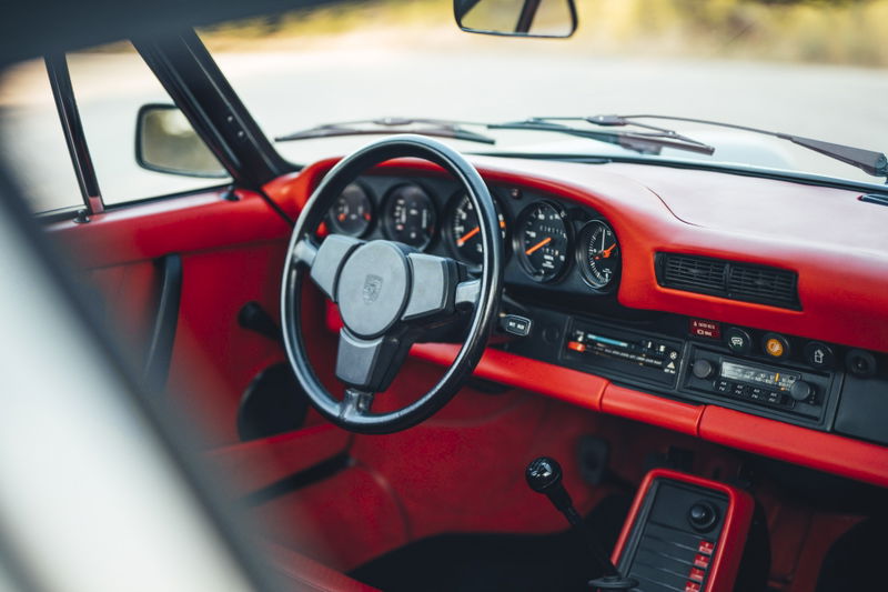 1977 Porsche 911S interior: iconic 5-dial dash, classic steering wheel, and vibrant red trim elements. Manual shifter visible.