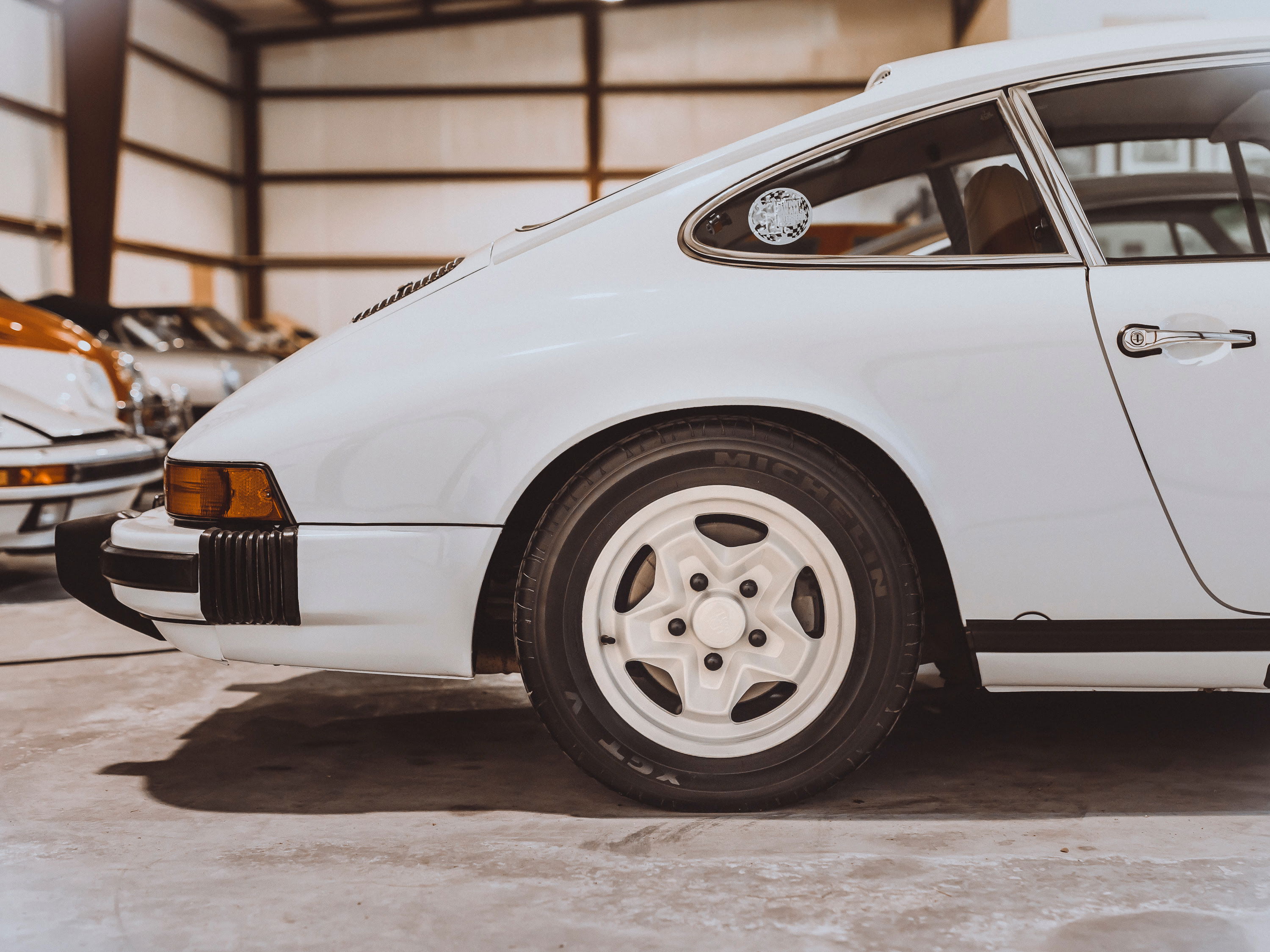 White vintage Porsche 911 G-series rear quarter view. Features white 5-spoke rims, black impact bumpers, chrome window trim, and handle.