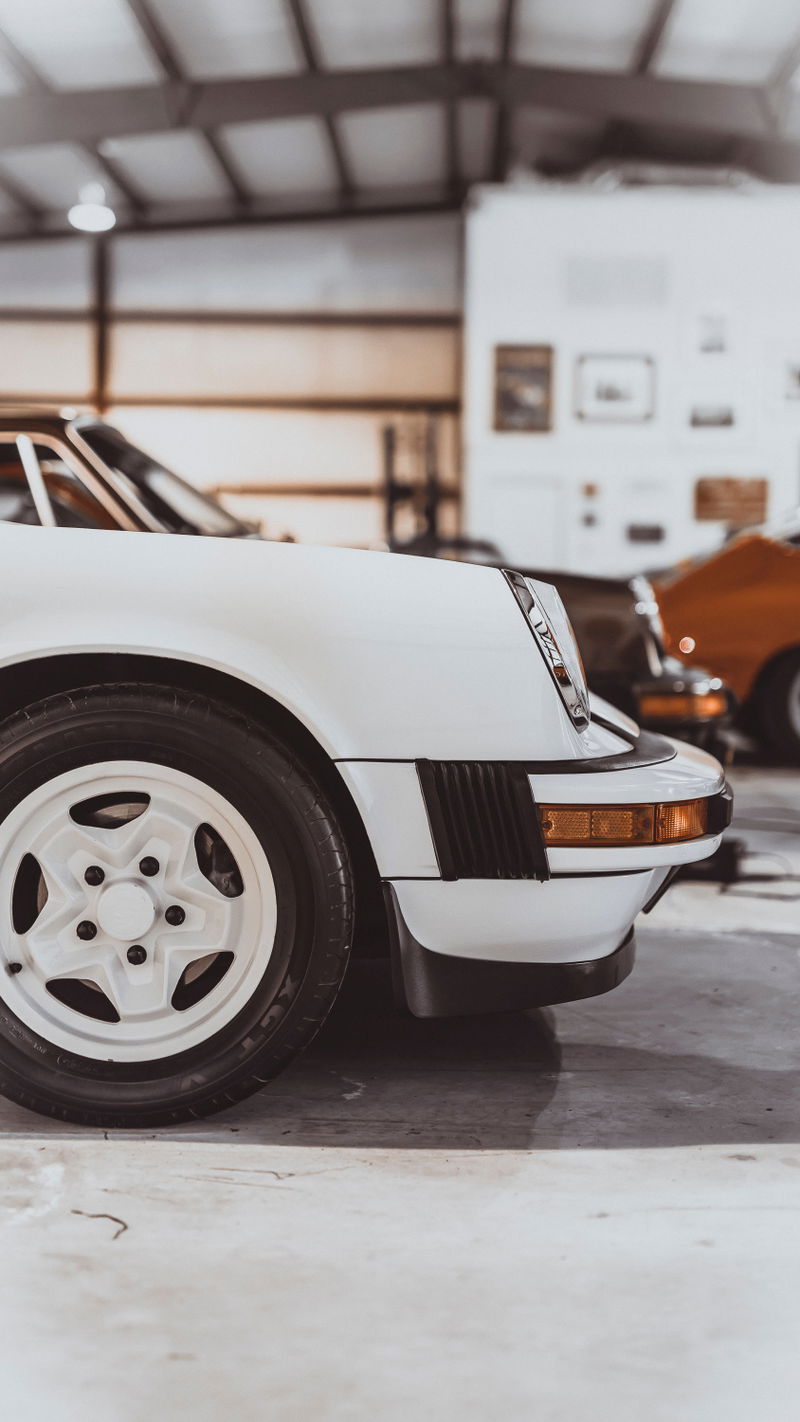 White Porsche 911 (G-series) front, featuring white 5-spoke wheels, black bumperettes, amber turn signal. Classic sports car.