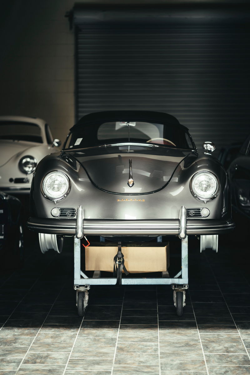 Front view of a metallic grey Porsche 356 Cabriolet on a workshop dolly, showcasing chrome bumpers, round headlights, and soft top.