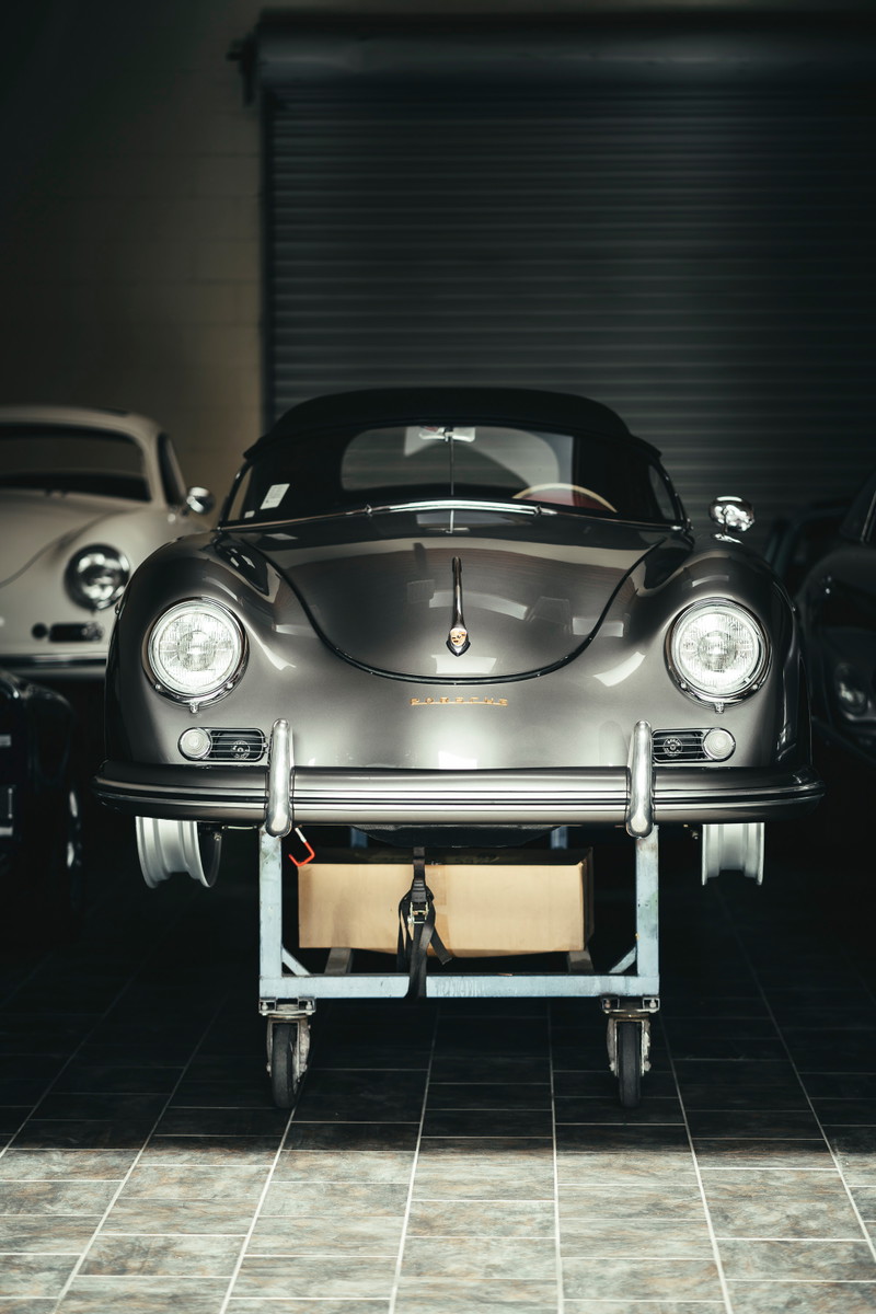 Front view of a metallic grey Porsche 356 Cabriolet on a workshop dolly, showcasing chrome bumpers, round headlights, and soft top.