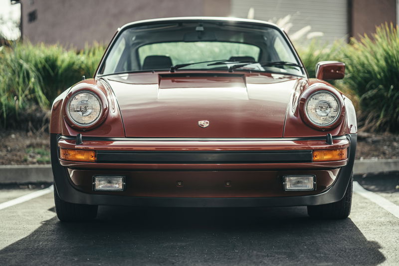 Front view of a deep red Porsche 911 G-series, showcasing its classic round headlights and US-spec rubber bumper trim.