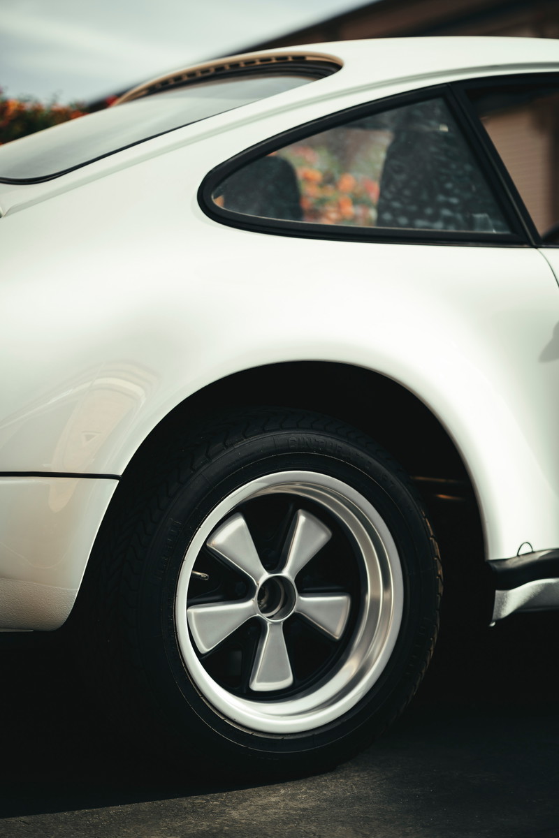 White classic Porsche 911 rear quarter panel, showcasing iconic polished Fuchs wheels, black window trim, and a sliding sunroof.