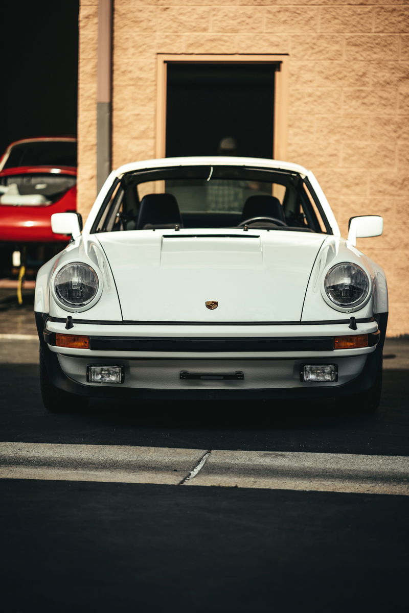 Frontal view of a pristine white classic Porsche 911 G-series with iconic round headlamps and black impact bumpers.