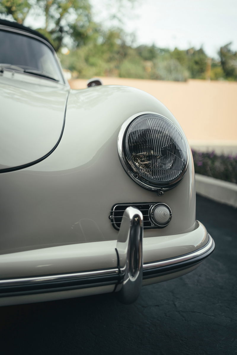 Close-up of a classic ivory Porsche 356 Cabriolet's front. Iconic round headlight, chrome trim, bumperette, and signal light.