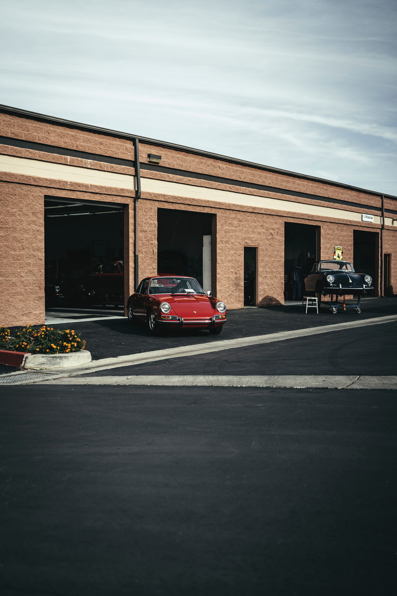 Vibrant red vintage Porsche 911, period-correct Fuchs-style wheels, parked outside a classic car restoration bay.