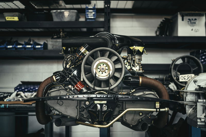 Restored Porsche 911 air-cooled flat-six engine with its iconic cooling fan and brown wrapped exhaust in a workshop setting.