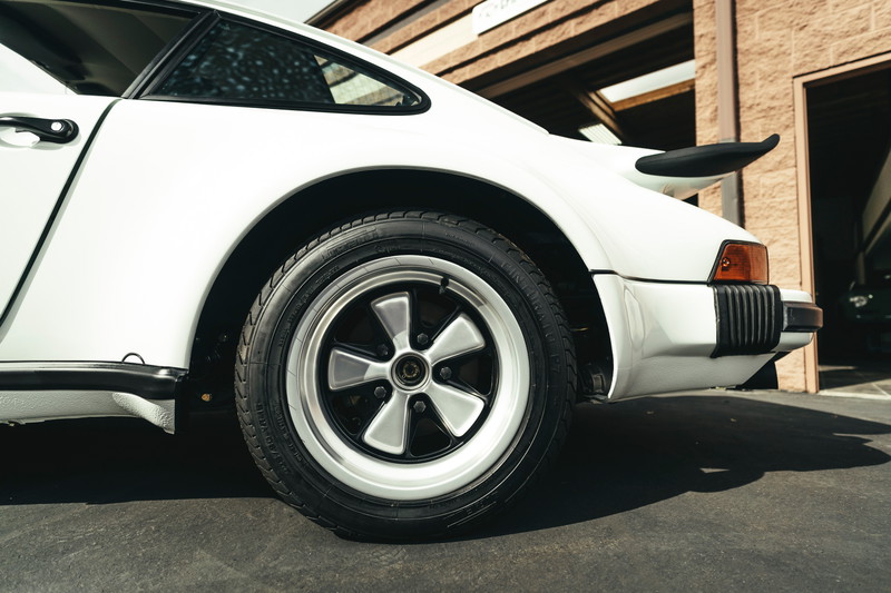White Porsche 911 G-series rear quarter, showcasing classic Fuchs rims, 'whale tail' spoiler, and black impact bumper.