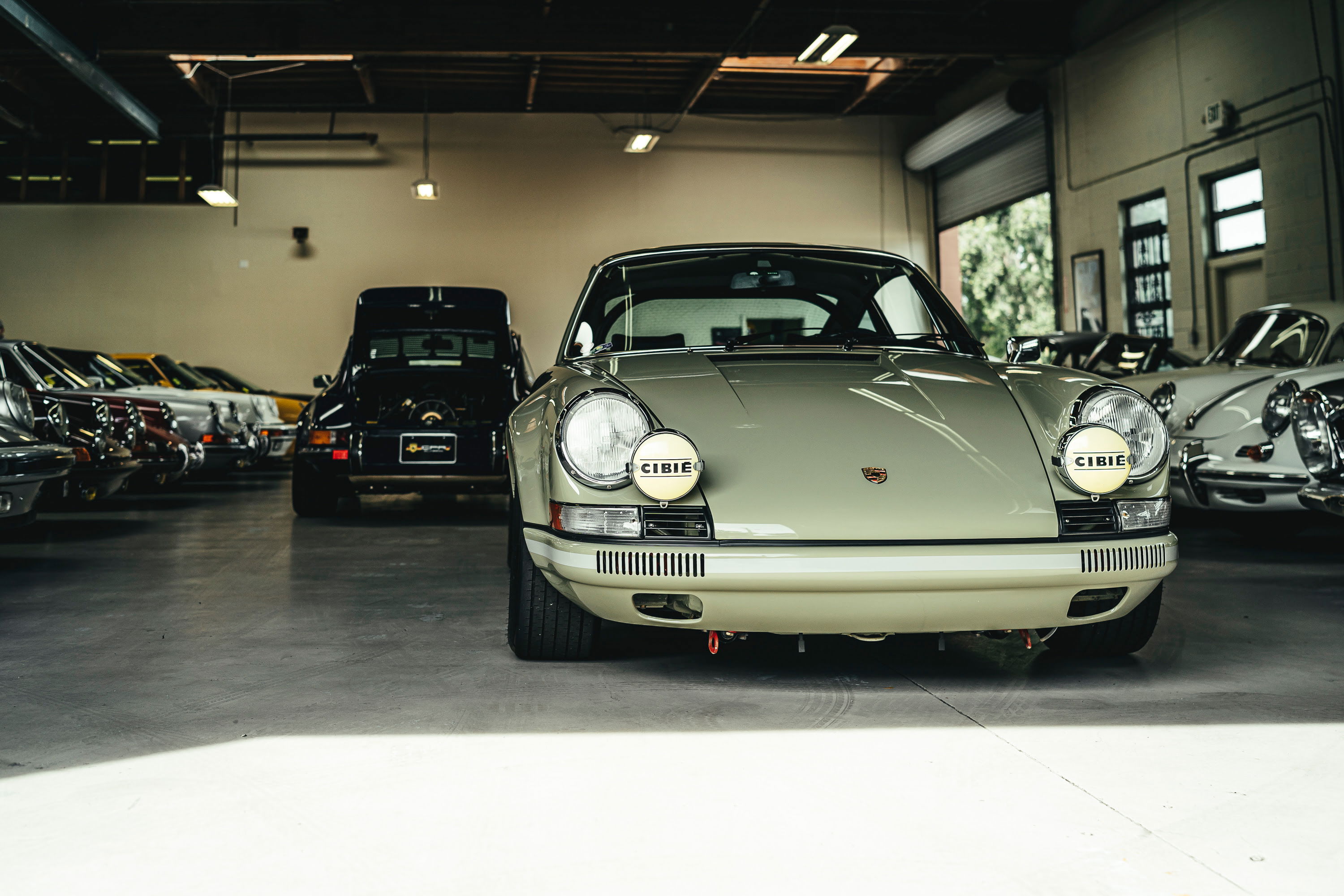 Sage green classic Porsche 911 sports car featuring prominent Cibie rally lights and a period-correct front bumper in a garage.