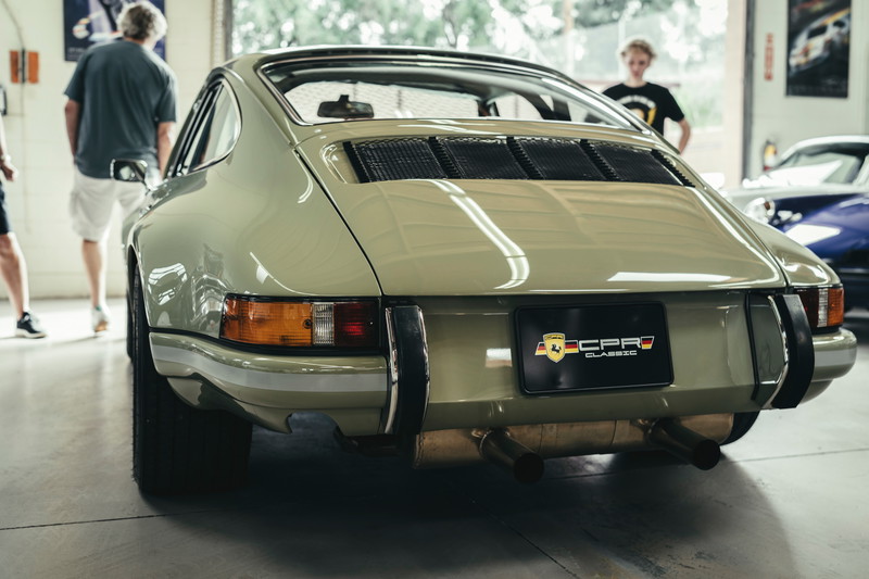 Vintage sage green Porsche 911 rear, featuring iconic engine lid grilles, chrome trim, bumperettes, and dual exhaust.