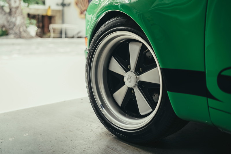 Close-up of a green Porsche 911's Fuchs-style alloy rims and tire, featuring a black side livery on the fender.