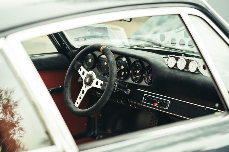 Interior of a classic air-cooled Porsche 911, showcasing its Alcantara steering wheel, five-gauge cluster, red trim, and rally timers.