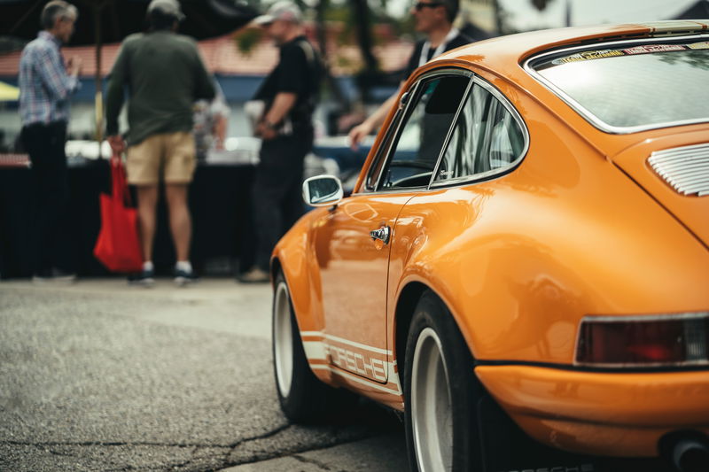 Tangerine Porsche 911 with white "PORSCHE" side livery, chrome trim, and white Fuchs-style rims. Lit Show LA.