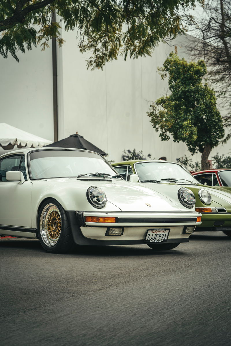 White Porsche 911 G-series coupe with gold multi-piece mesh rims and black bumper trim, parked with other classic 911s.