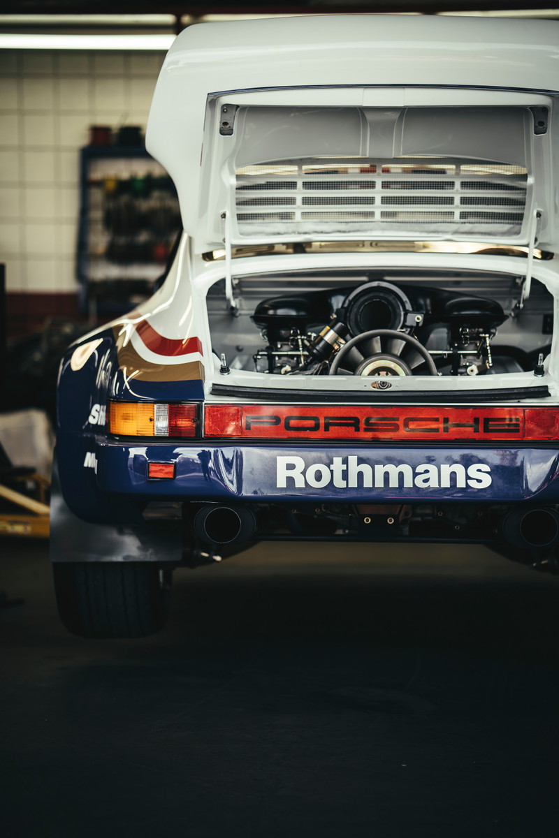 Classic Porsche 911 rally car, white with Rothmans livery. Open decklid exposes its flat-six engine. Rear view shows mud flaps & dual exhausts.