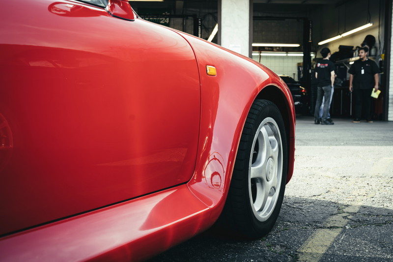 Close-up of a vibrant red Honda S2000's front fender, amber side marker, and silver 5-spoke AP1 OEM rims in a garage.