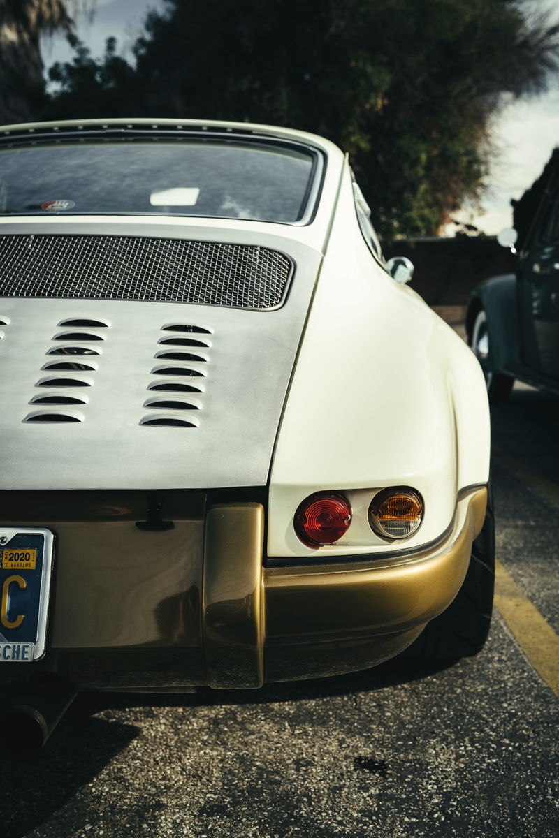 Rear view of a classic two-tone Porsche 911, off-white with bronze lower bumper & trim. Engine lid features a mesh grille and horizontal louvers.
