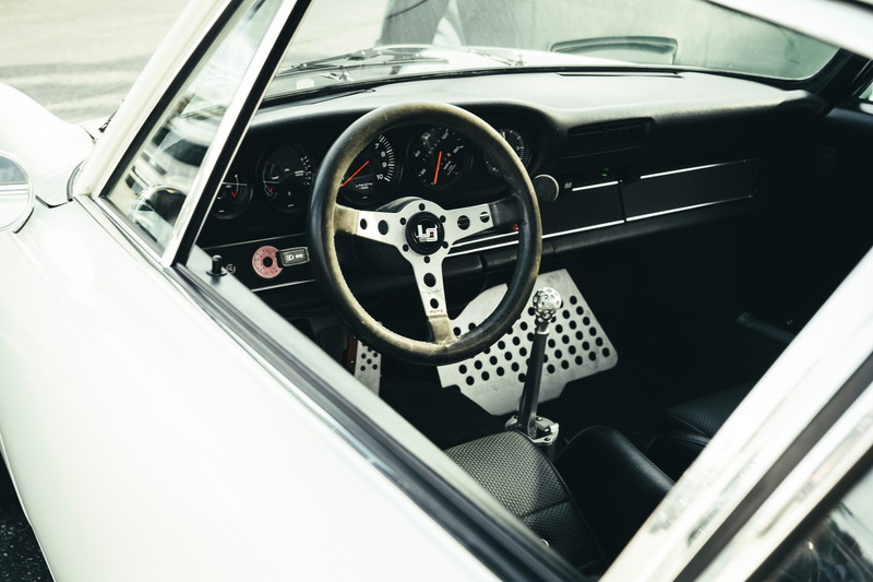 Classic white Porsche 911 interior: aftermarket racing steering wheel, perforated aluminum pedals, and manual shifter.