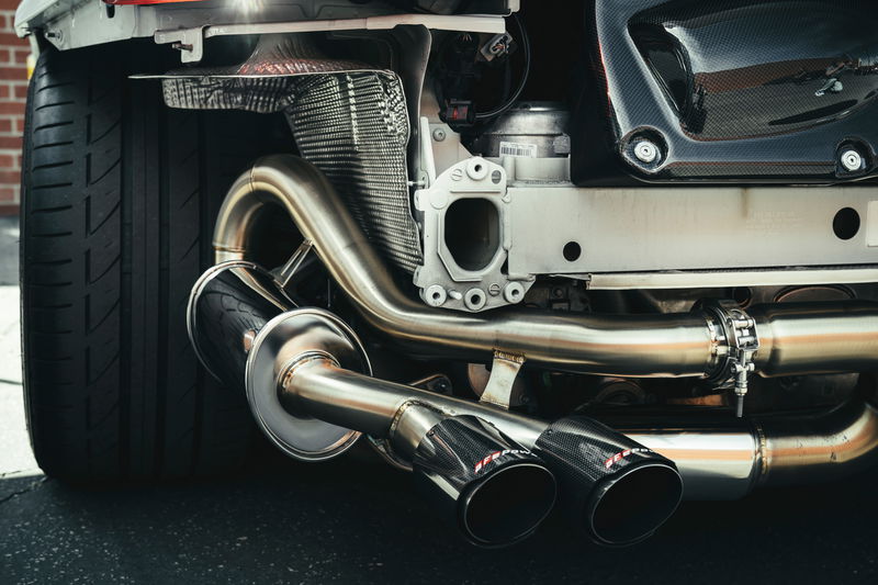 White performance car rear quarter: Akrapovič polished stainless steel exhaust, dual carbon fiber tips, exposed chassis, wide tire.
