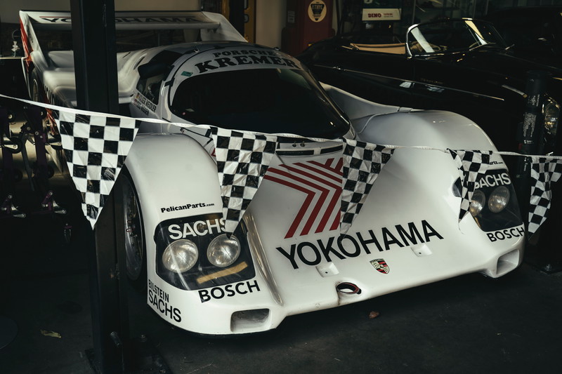 Iconic white Porsche 962C Kremer Group C prototype, featuring YOKOHAMA/SACHS livery and exposed racing rims, draped with flags.