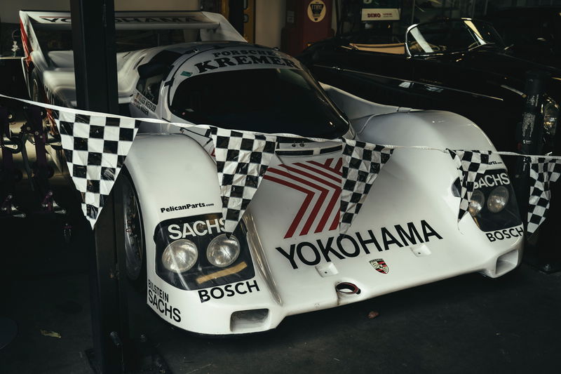 Iconic white Porsche 962C Kremer Group C prototype, featuring YOKOHAMA/SACHS livery and exposed racing rims, draped with flags.