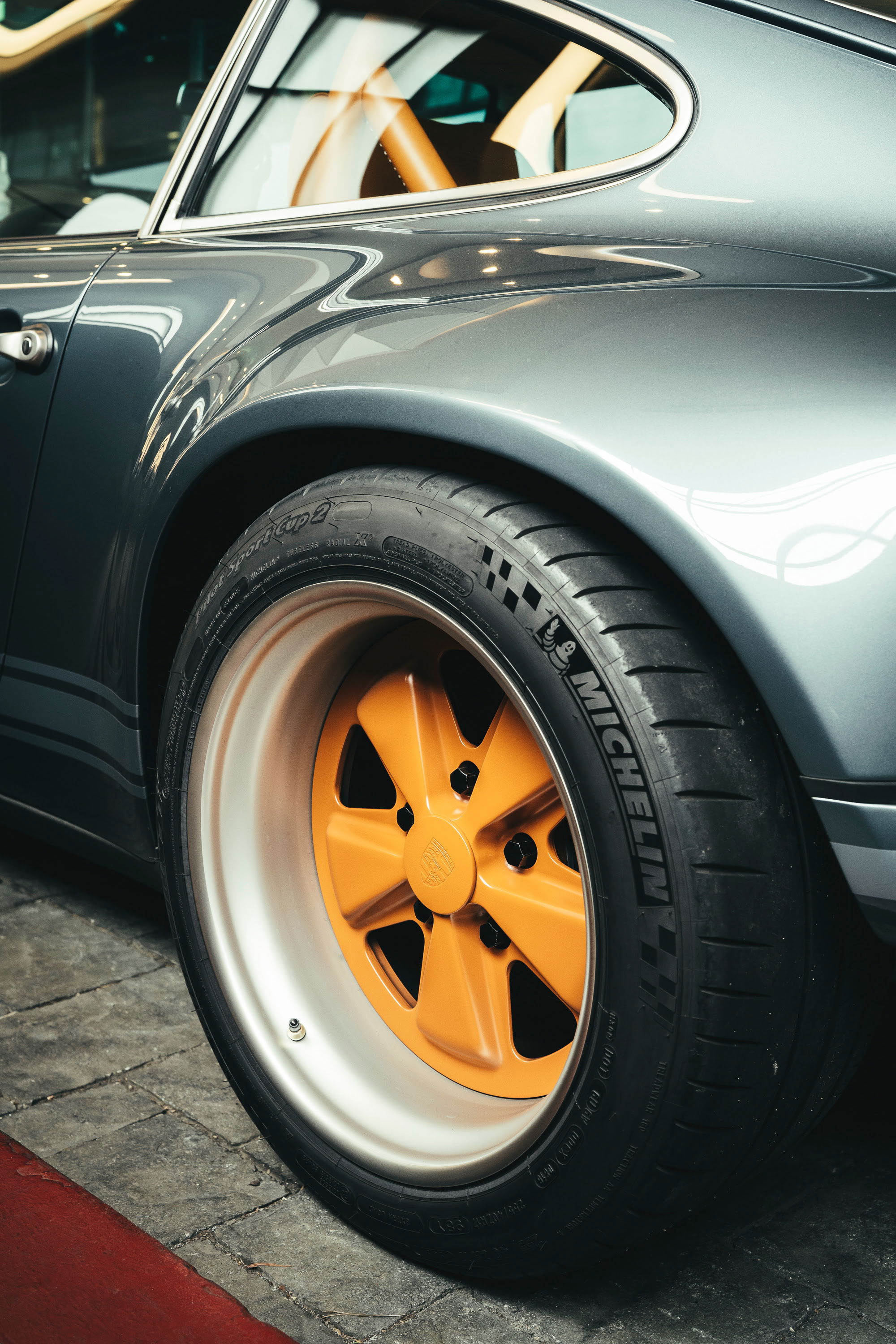 Grey air-cooled Porsche 911 restomod, detailed with custom orange Fuchs-style rims, Michelin Pilot Sport Cup 2 tires, and tan interior.