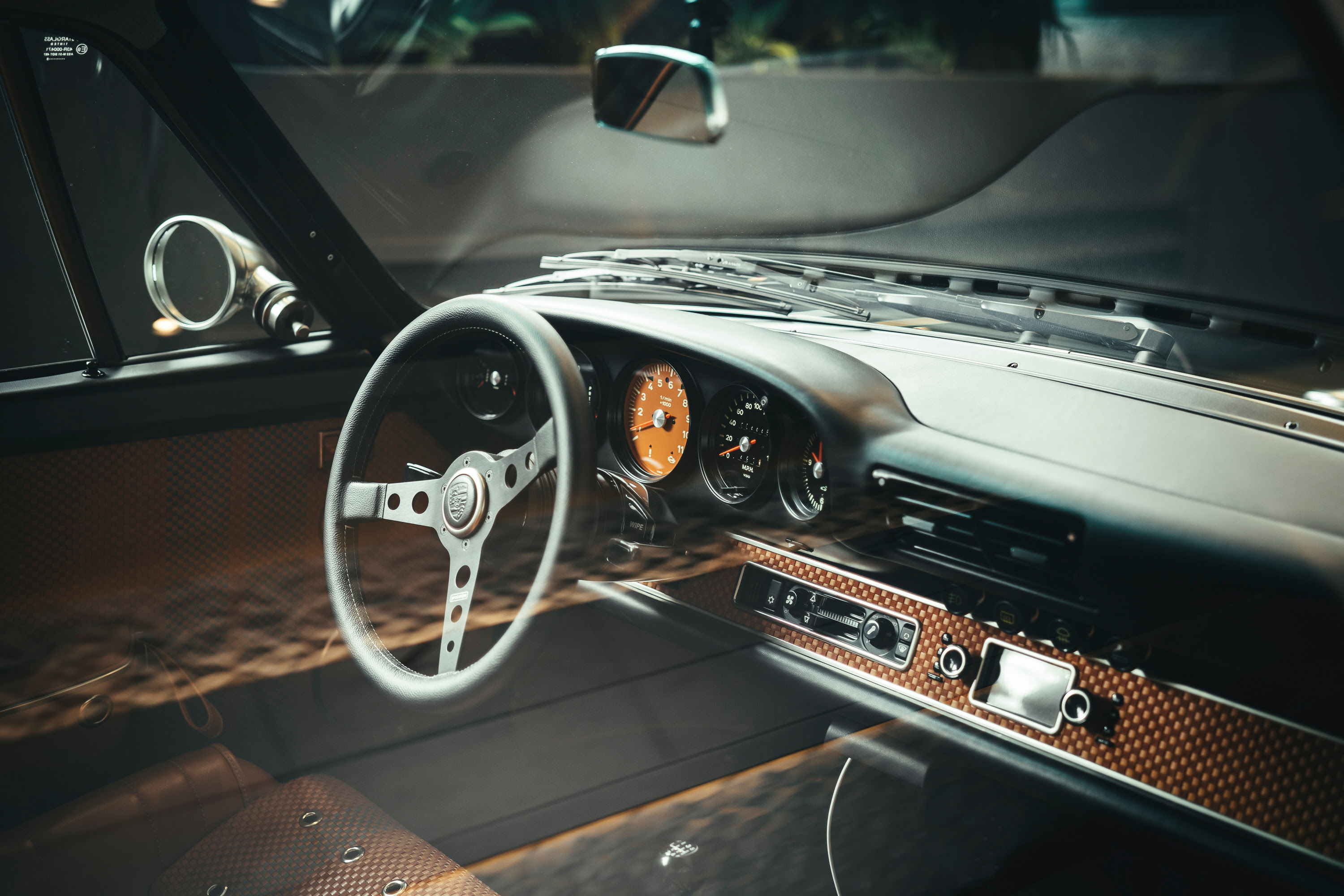 Detailed interior of a classic air-cooled Porsche 911, featuring a perforated three-spoke steering wheel, orange-faced gauges, and woven dash trim.