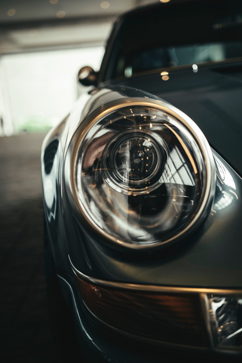 Close-up of a classic Slate Grey Porsche 911 headlight, highlighting its iconic round design, polished chrome bezel, and amber turn signal.