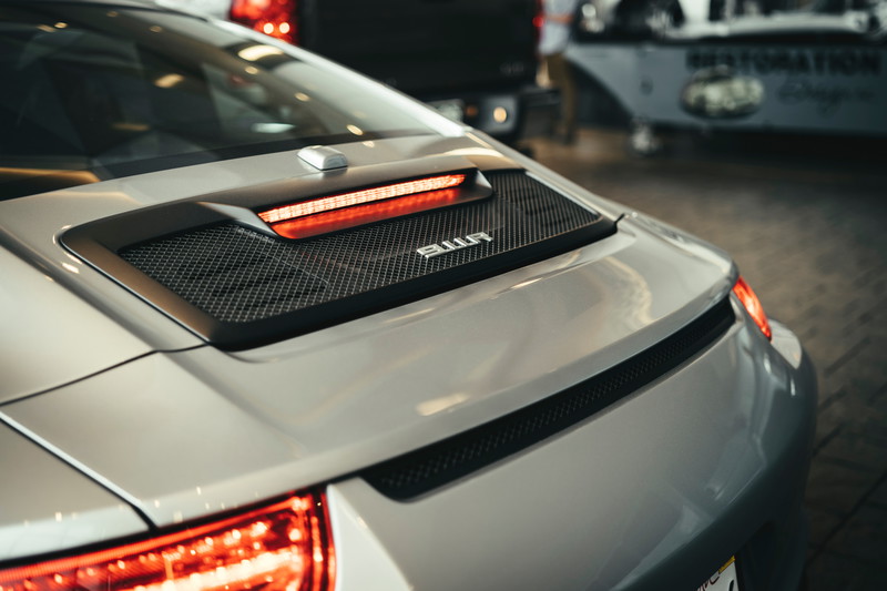 Silver Porsche 992 911 rear view, showcasing its illuminated LED taillight strip, engine cover's mesh grille, and 911 badge.