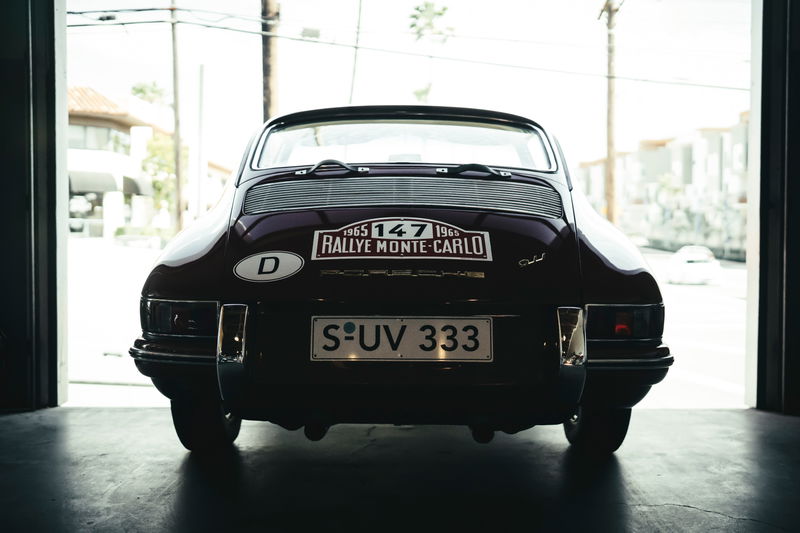 Rear view of a dark red 1965 Porsche 911, showcasing Monte Carlo Rallye livery, chrome bumperettes, and German plates.