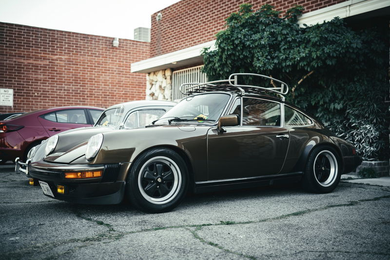 Vintage dark brown Porsche 911 with Fuchs-style rims, widebody flares, and period roof rack at a Los Angeles car show.