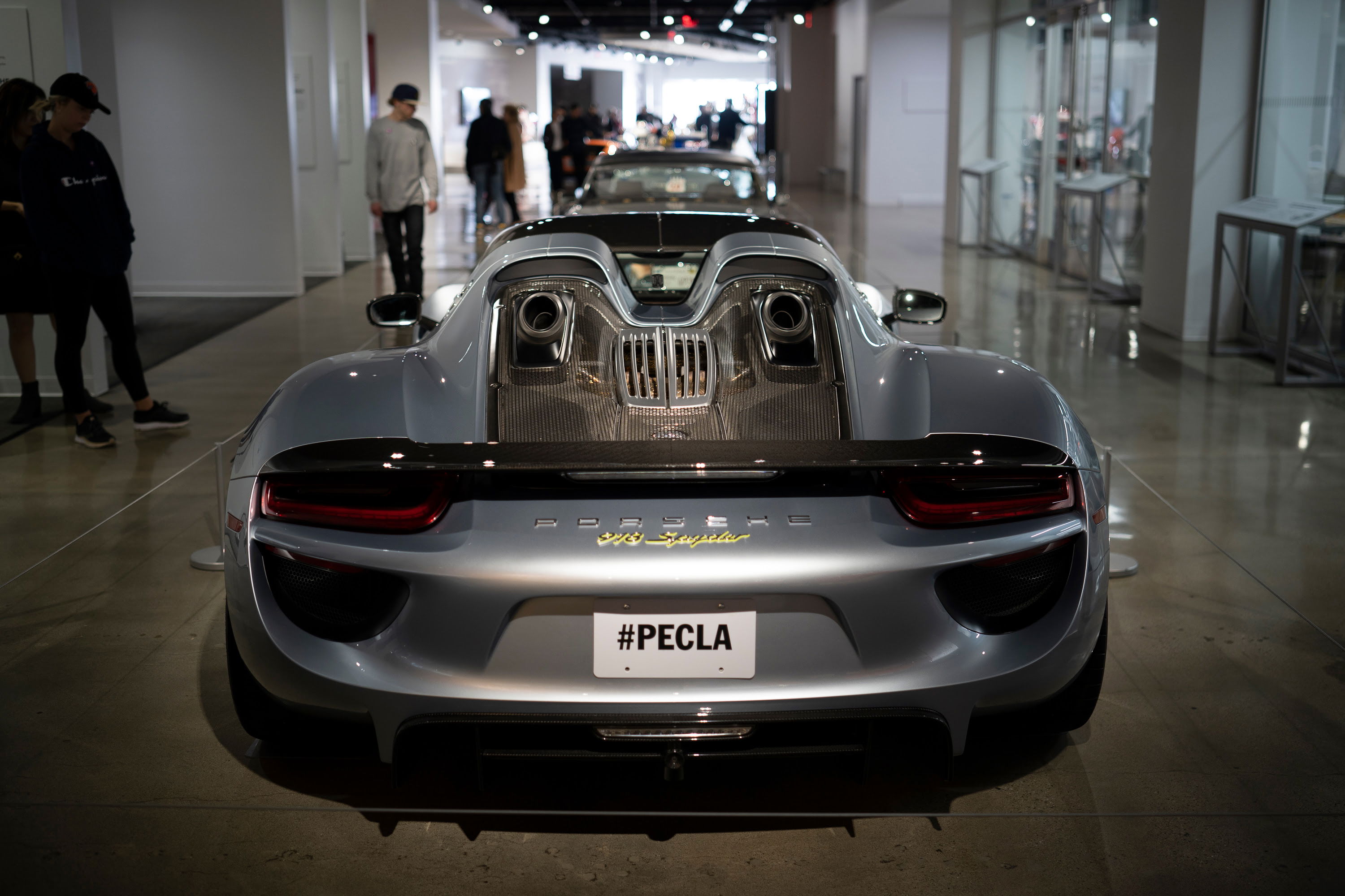Metallic silver Porsche 918 Spyder rear view, featuring top-exit exhausts, carbon fiber trim, and gold badging. At PECLA.