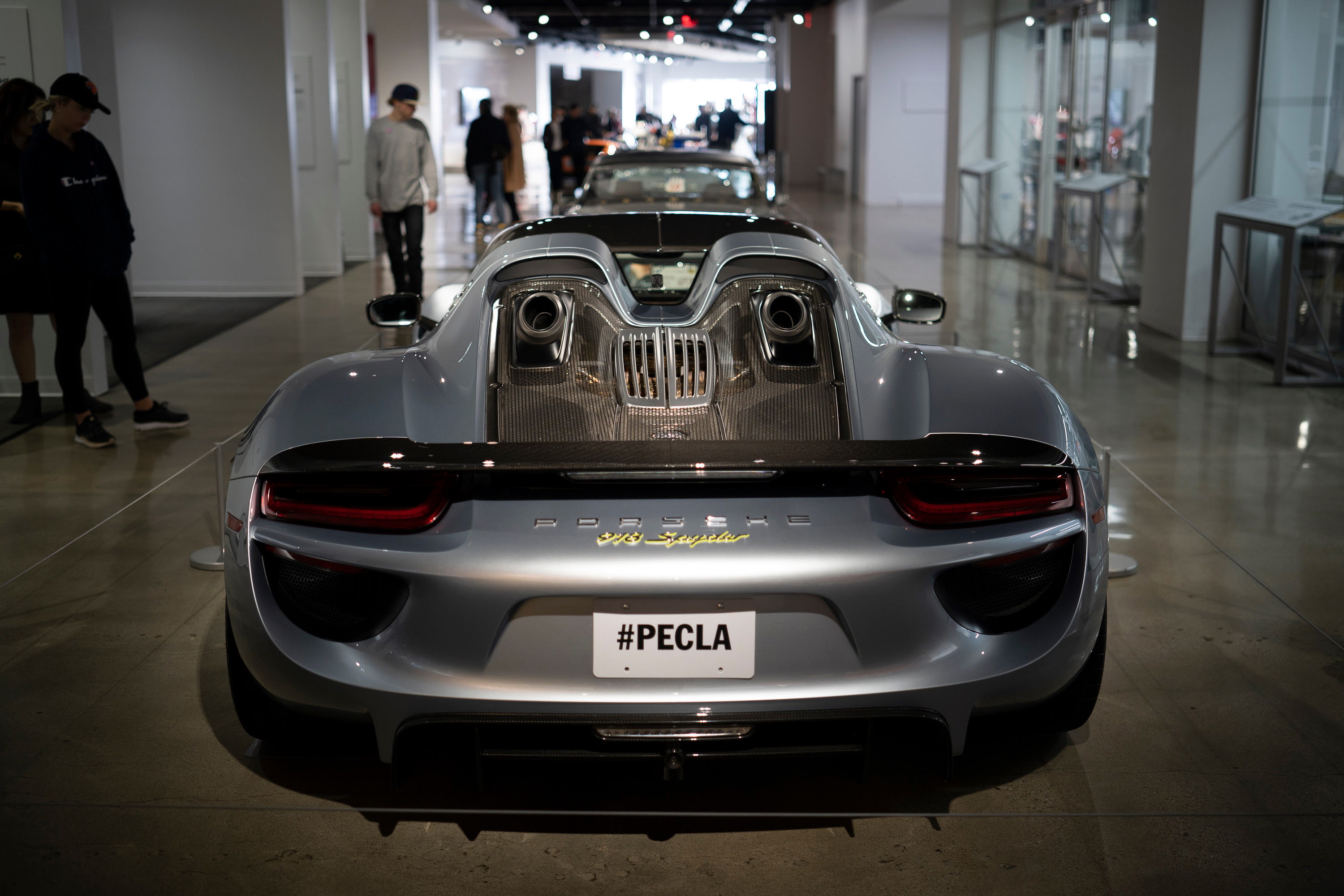 Metallic silver Porsche 918 Spyder rear view, featuring top-exit exhausts, carbon fiber trim, and gold badging. At PECLA.