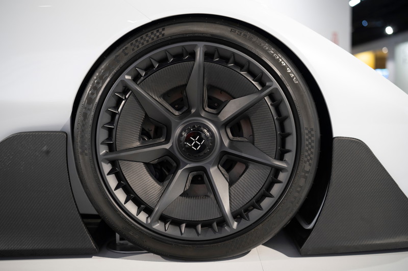 Close-up of a white Deus Vayanne electric hypercar's front wheel. Features dark multi-spoke rims, performance tires, & carbon fiber aero trim.