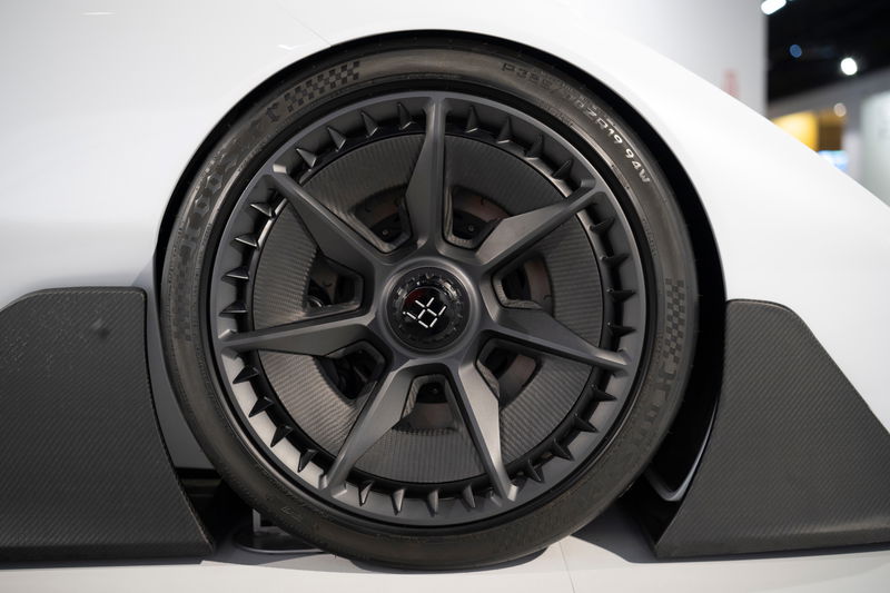 Close-up of a white Deus Vayanne electric hypercar's front wheel. Features dark multi-spoke rims, performance tires, & carbon fiber aero trim.