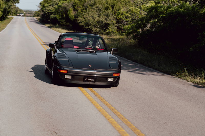 Black 1989 Porsche 930 Turbo Flachbau with red interior drives on a country road.