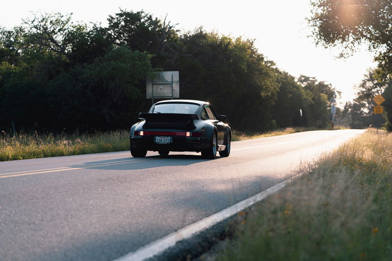Black 1989 Porsche 930 Turbo Flachbau driving on a road with a rear spoiler.