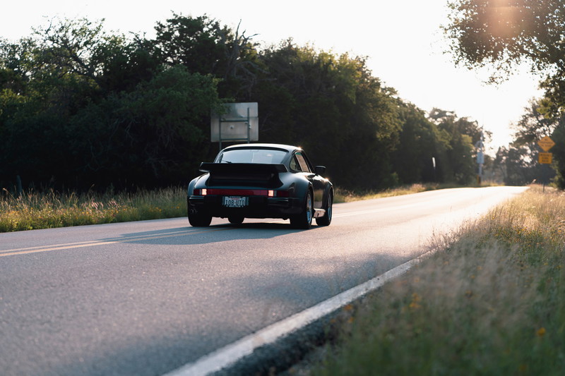 Black 1989 Porsche 930 Turbo Flachbau driving on a road with a rear spoiler.