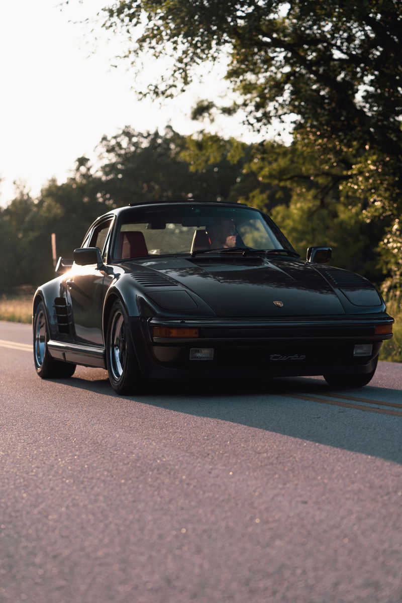 Black 1989 Porsche 930 Turbo Flachbau on road, showing its wide body kit and signature rims.