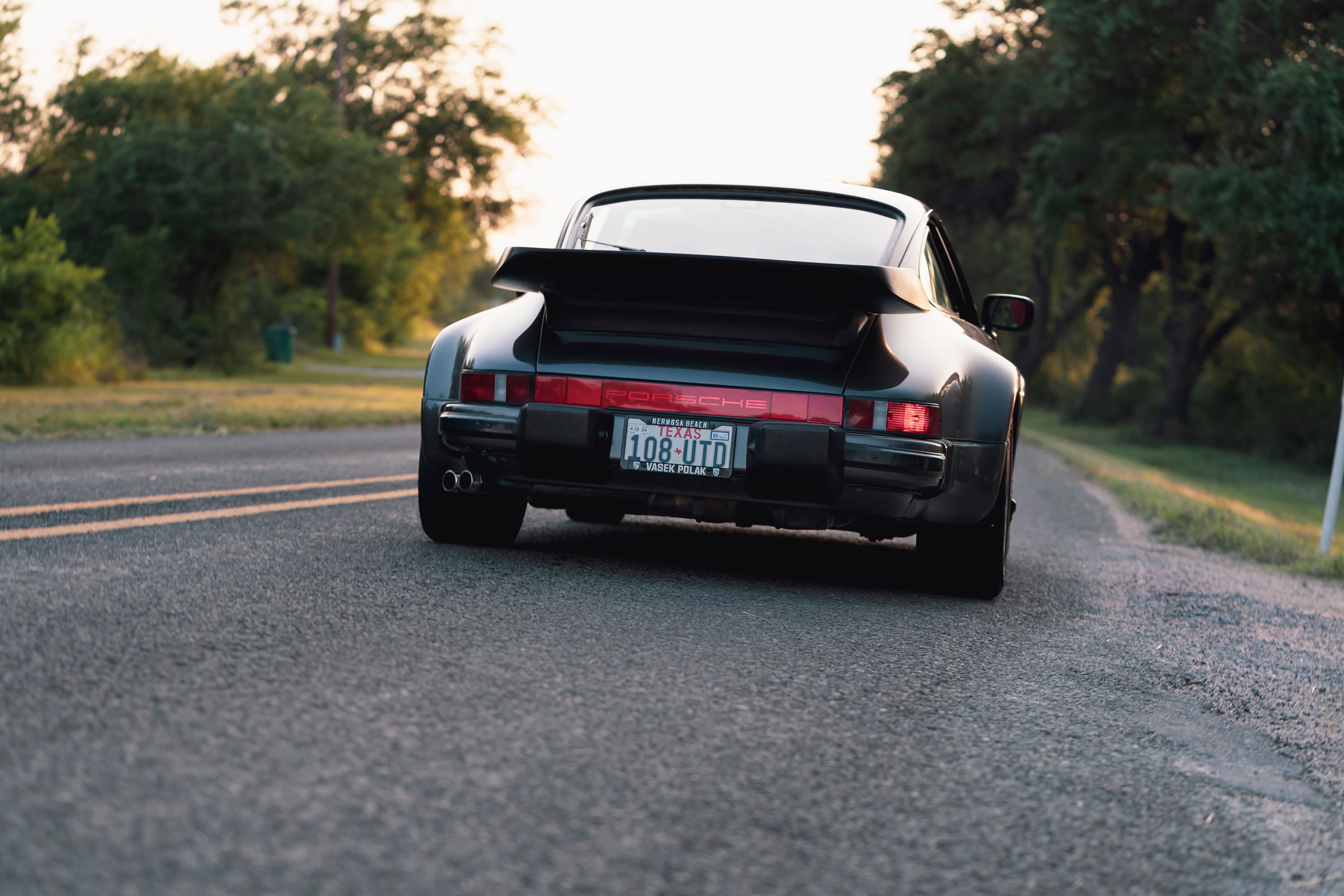 Rear view of a black 1989 Porsche 930 Turbo Flachbau with a large spoiler on a rural road.