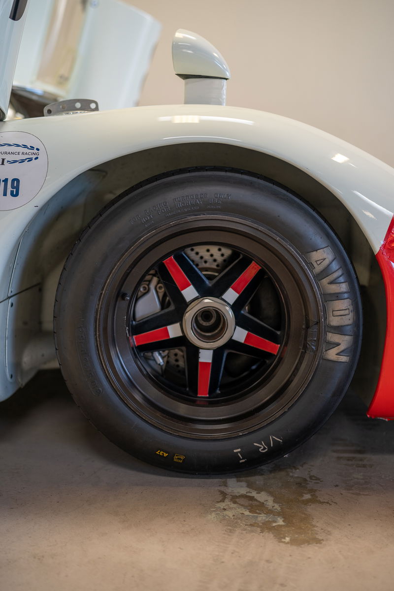 Close-up of a white Porsche 911 race car's wheel, featuring a black 5-star rim with red and silver accents, and Avon tires.