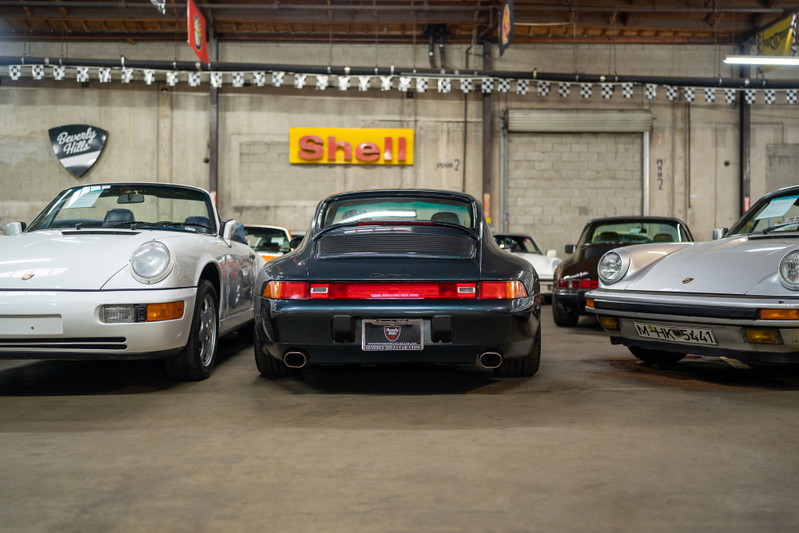 Rear view of a dark green Porsche 911 Carrera in a showroom, with other Porsches visible.