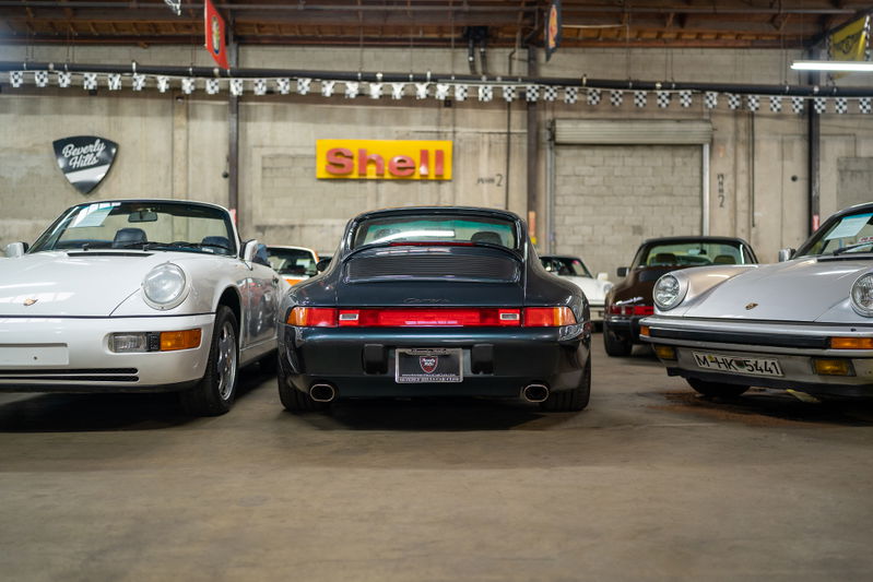 Rear view of a dark green Porsche 911 Carrera in a showroom, with other Porsches visible.