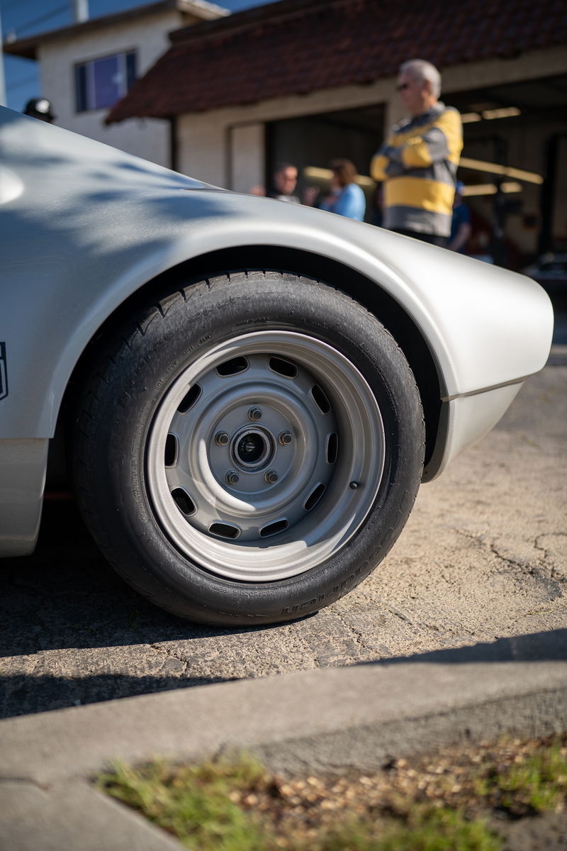 Close-up of a silver vintage race car's rear wheel and fender. The classic steel rim has slotted holes.