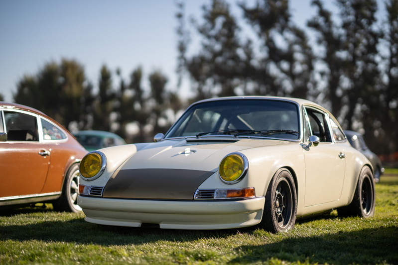 Vintage Porsche 911 with yellow headlights, custom trim, and black rims parked on grass at a car show.