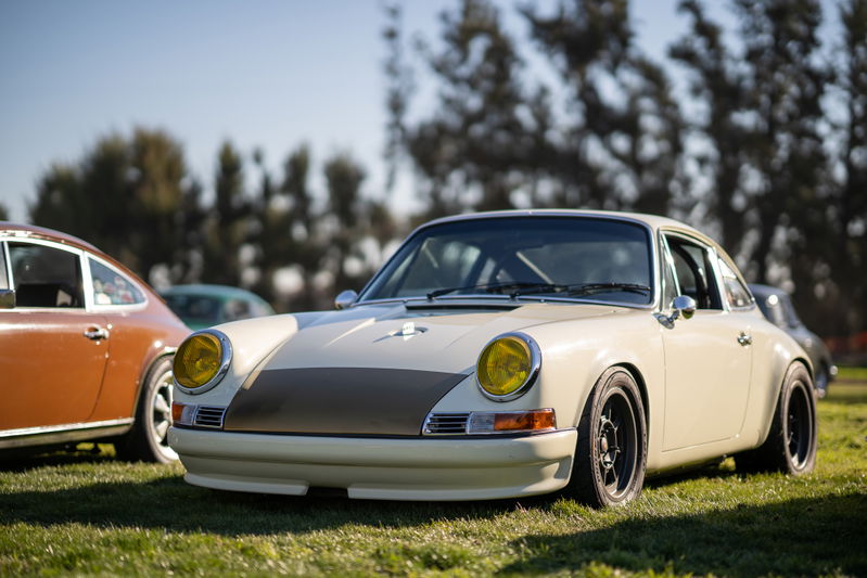 Vintage Porsche 911 with yellow headlights, custom trim, and black rims parked on grass at a car show.
