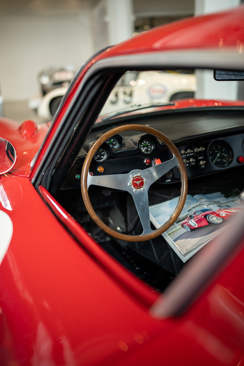 Close-up interior of a red vintage race car, showcasing a wooden steering wheel with a polished metal hub and emblem.