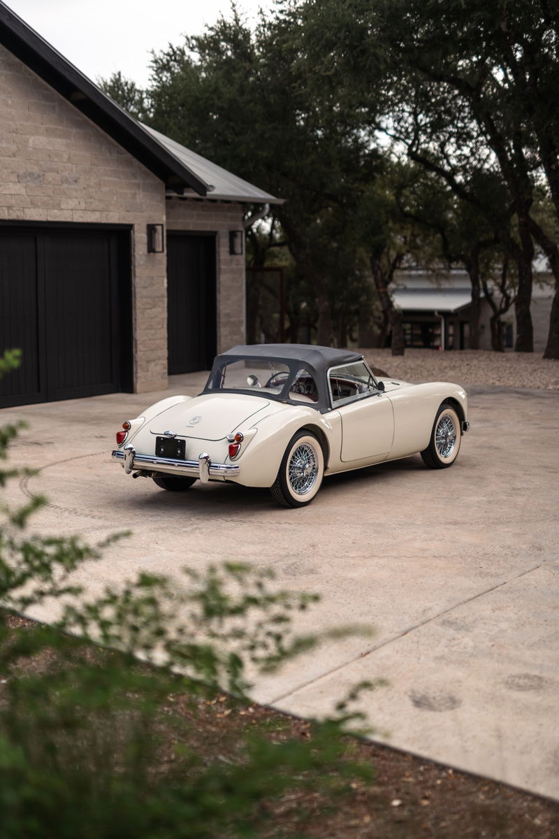 Rear 3/4 view of a cream 1957 MGA Roadster, with black soft top, wire wheels, and chrome trim.